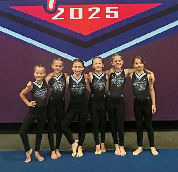 Six girls from North Carolina’s gymnastics team pose in front of a 2025 championship backdrop.
