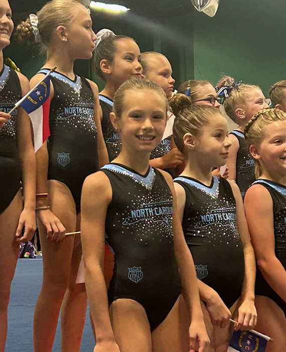Team North Carolina gymnasts stand smiling in leotards at a competition representing their state.