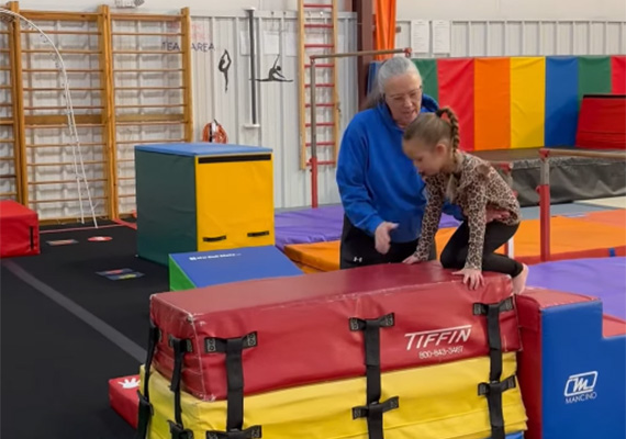 adc6 Coach assists a child climbing on stacked gymnastics mats during a preschool gymnastics class.