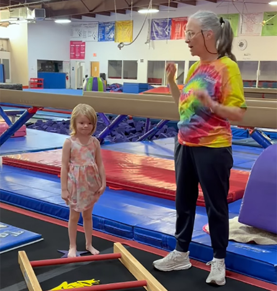 Gymnastics coach instructs a young girl standing beside low bars in a colorful training gym.