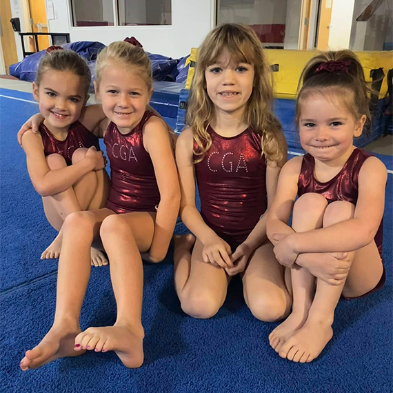 ss2 Four young girls in maroon CGA leotards sit smiling on the blue mat before class begins.