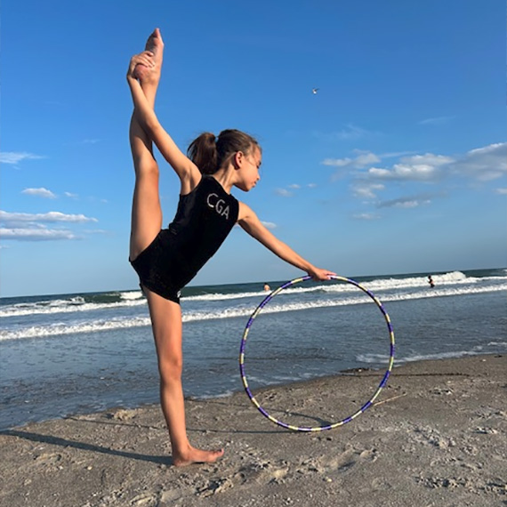 CGA gymnast practices rhythmic gymnastics on the beach, balancing in a pose with a hoop in hand by the ocean.